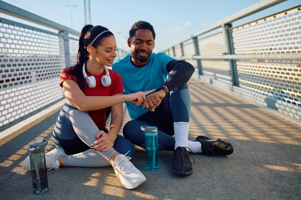 Smiling sports couple using fitness tracker while relaxing outdoors.